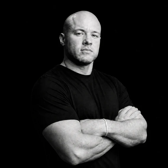 Black-and-white studio portrait of a man with a shaved head in a black T-shirt, arms crossed, looking at the camera against a black background.